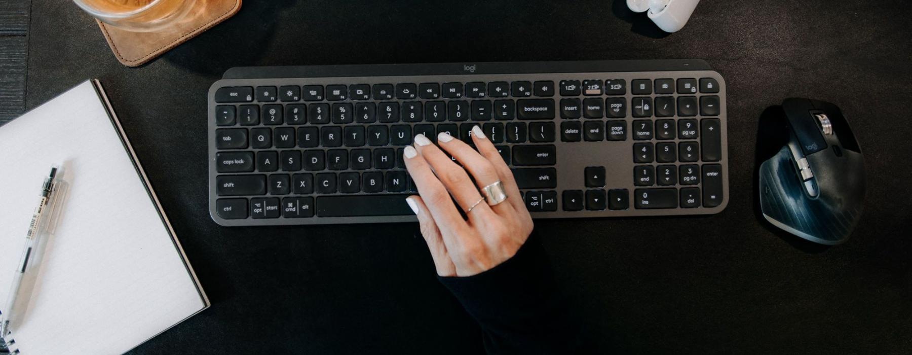 woman's hand on a keyboard surrounded by office items and a cup of tea on a coaster
