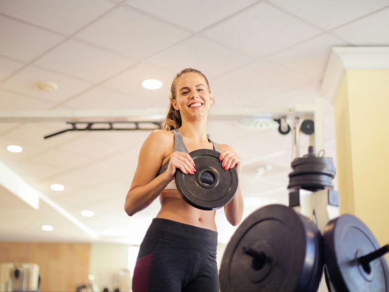 Montaire Student Living Apartments Smiling woman in workout clothes holding a weight plate at a gym, with gym equipment in the foreground.