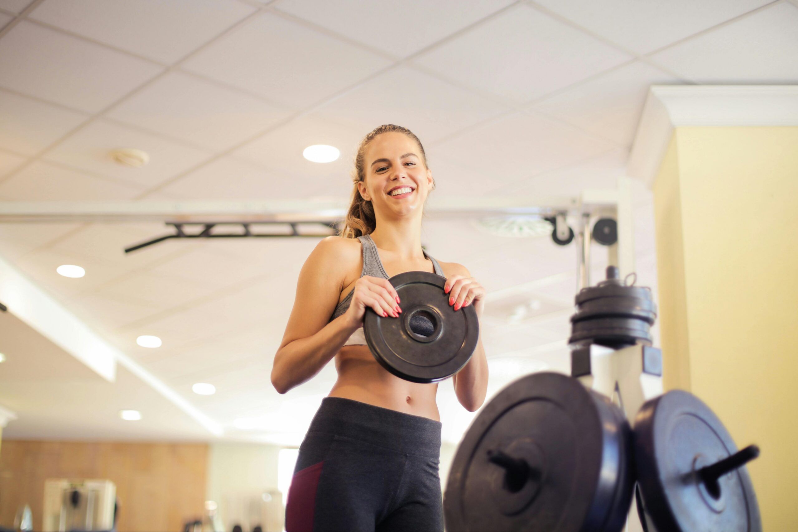 Montaire Student Living Apartments Smiling woman in workout clothes holding a weight plate at a gym, with gym equipment in the foreground.