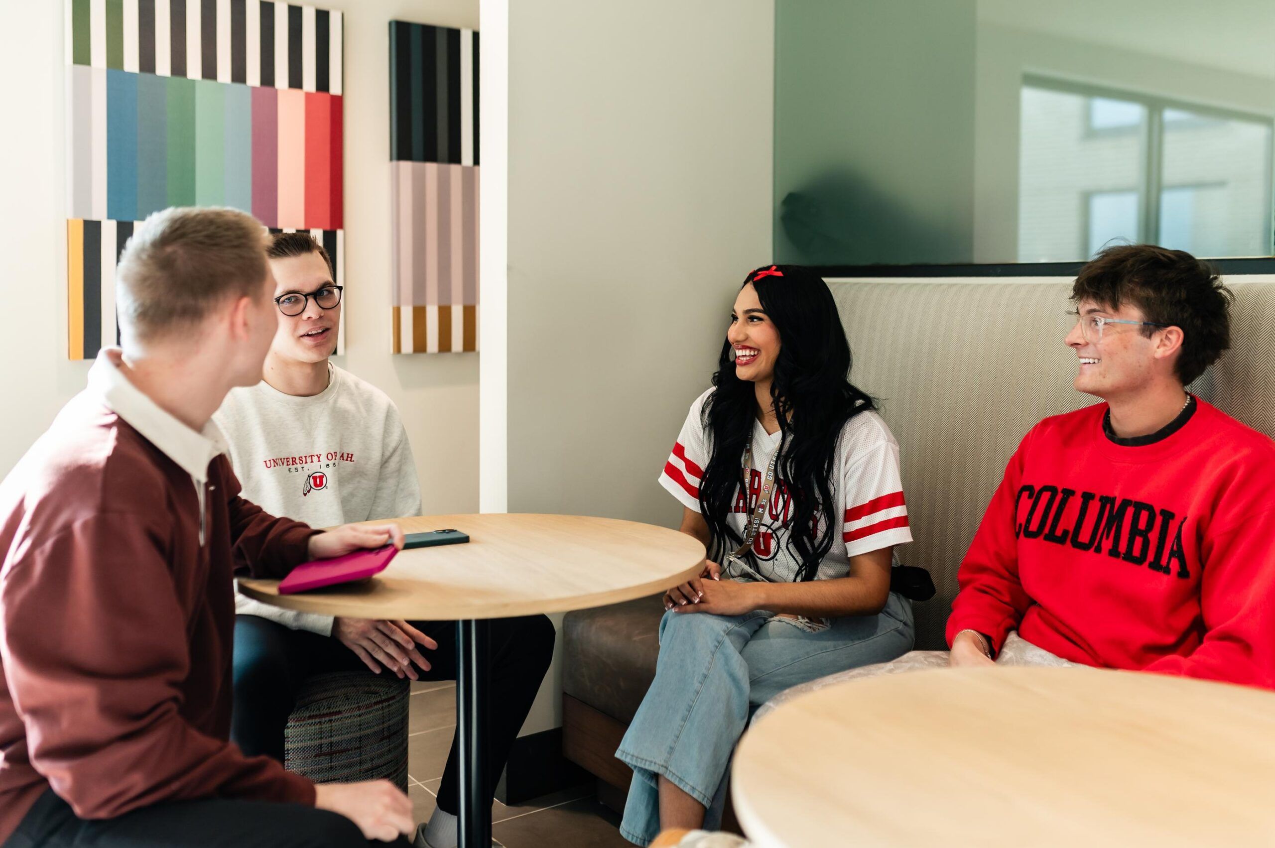 Montaire Student Living Apartments Four college students sit and talk around tables in a bright room, smiling and wearing university shirts.