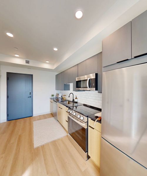 Montaire Student Living Apartments Modern kitchen with stainless steel appliances, wood flooring, and gray cabinets, viewed from the entrance.