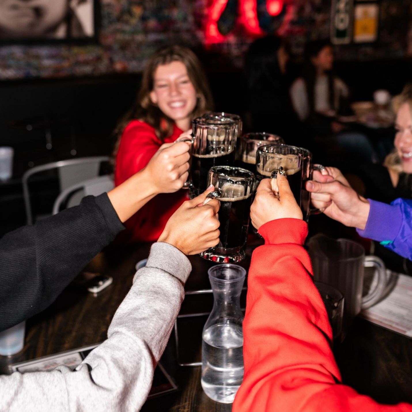 Montaire Student Living Apartments Four friends clink beer mugs at a lively bar with a glowing Pi symbol on the wall behind them.