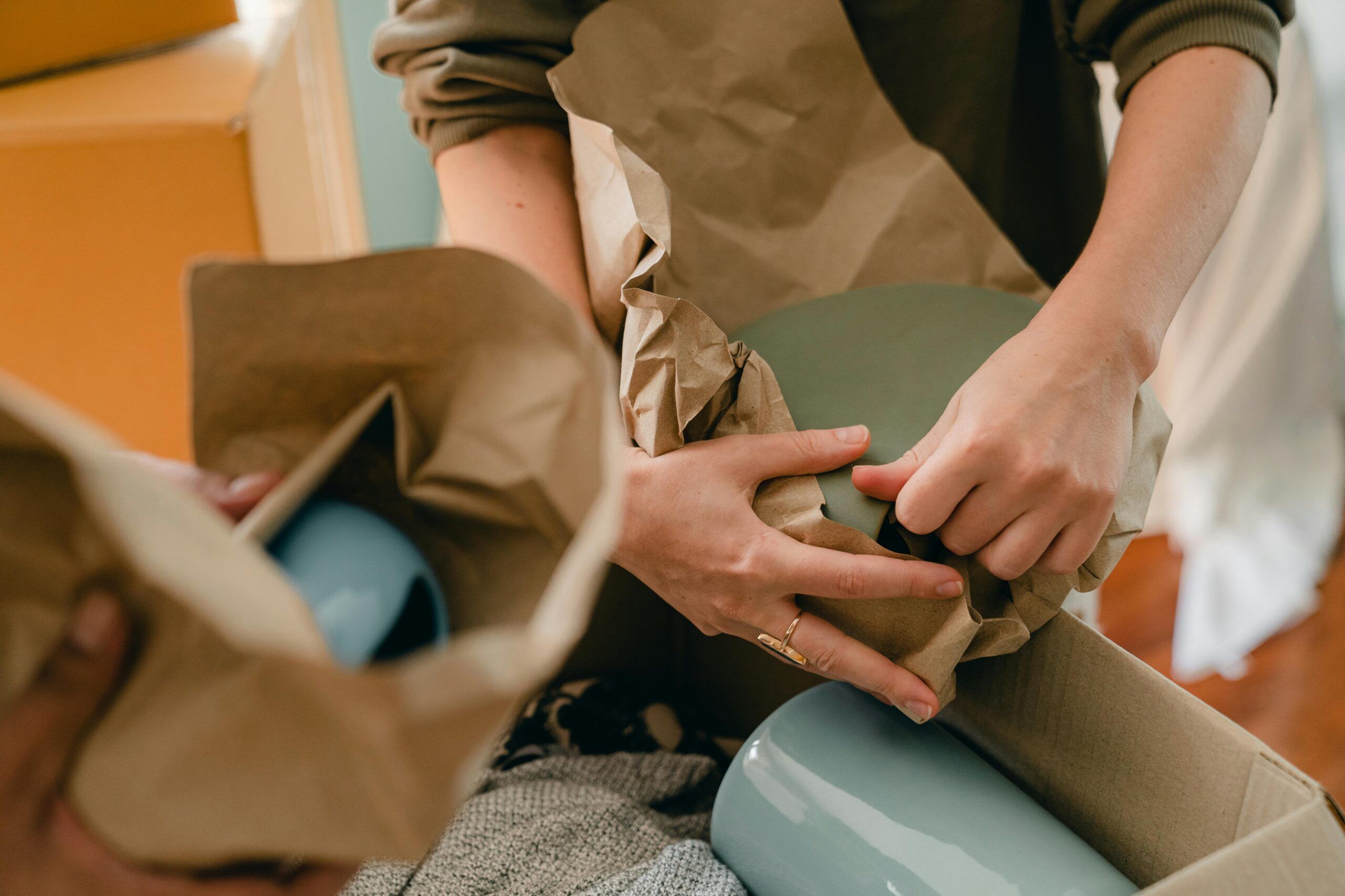 Montaire Student Living Apartments Hands wrapping blue ceramics in brown paper for packing, with boxes and cloth visible in the background.