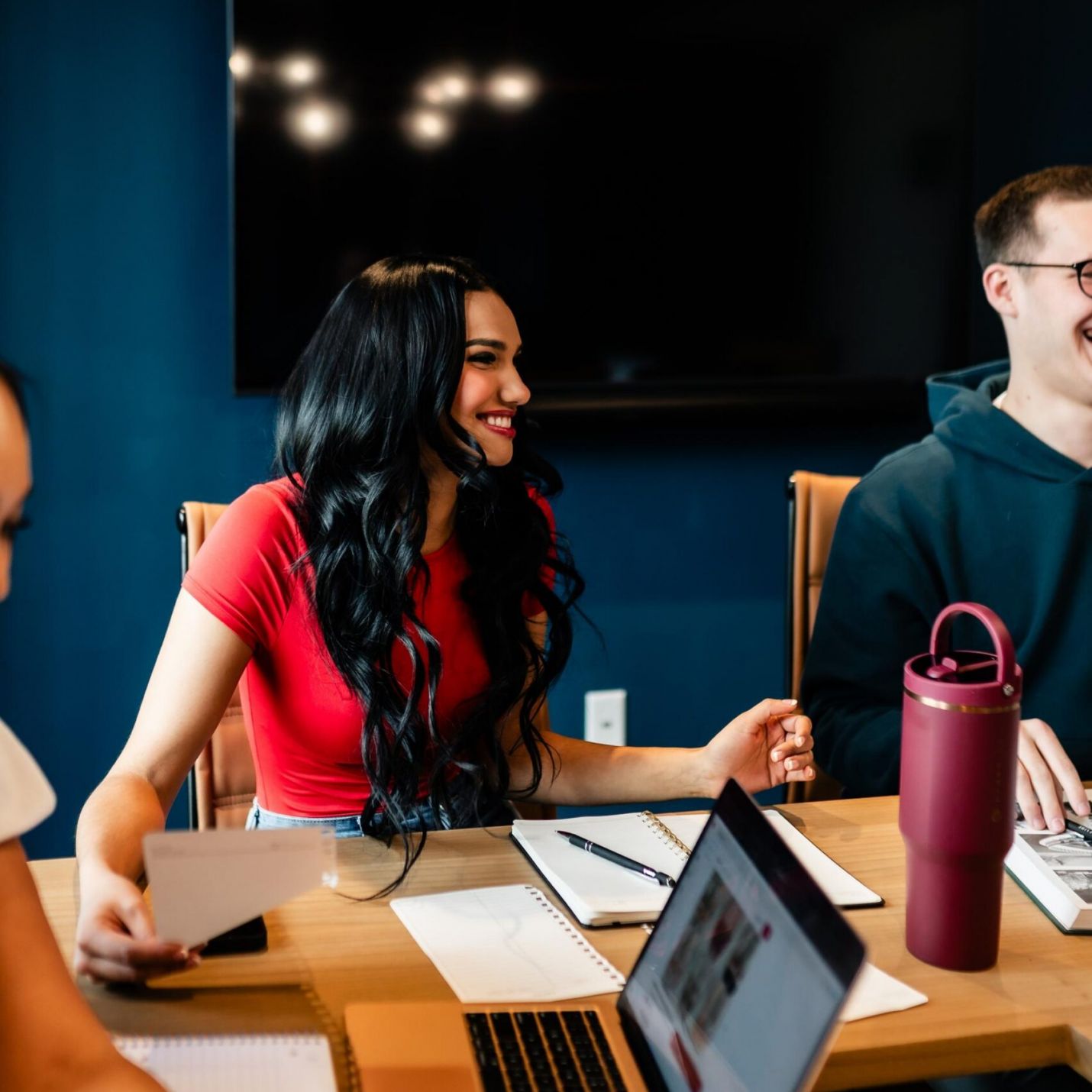 Montaire Student Living Apartments Woman in a red shirt smiles while sitting at a table with a laptop, notebooks, and water bottle.