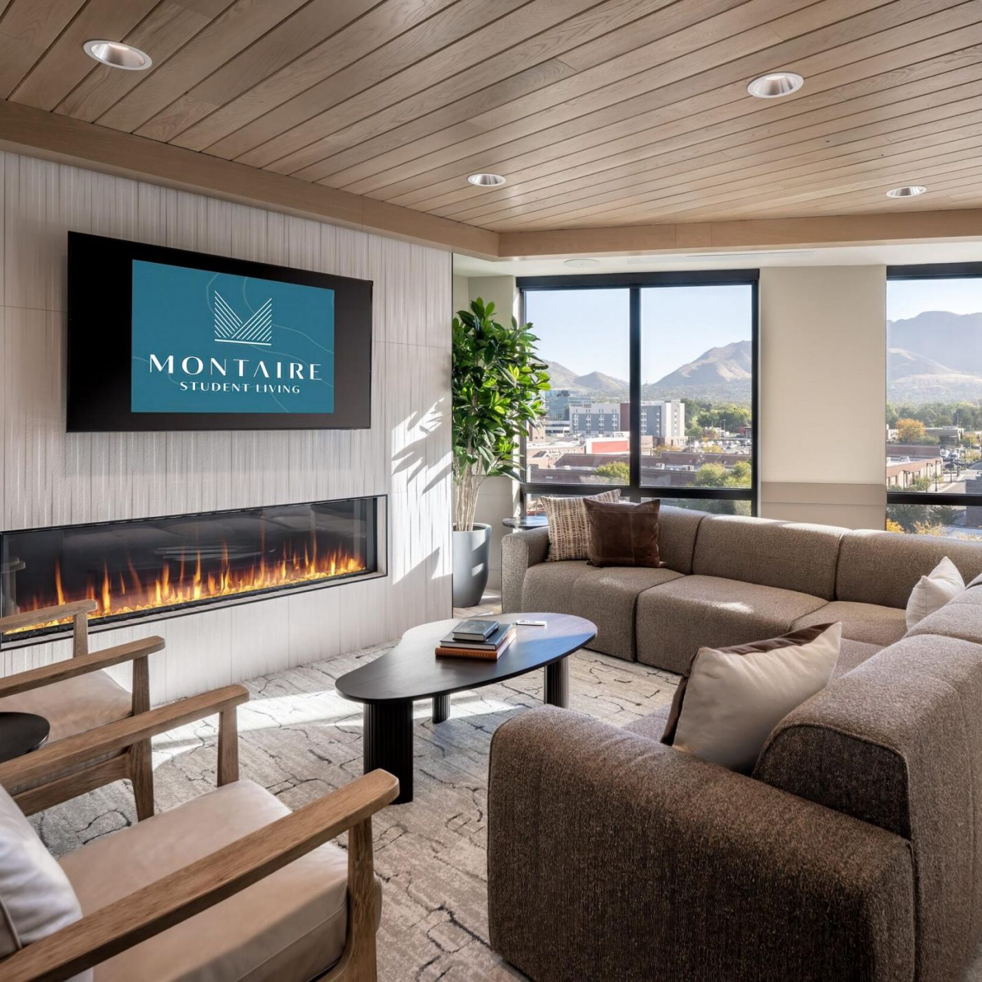 Montaire Student Living Apartments A lounge with a sectional sofa, armchairs, and a fireplace beneath a television displaying the Montaire Student Living logo.