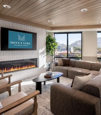 Montaire Student Living Apartments A lounge with a sectional sofa, armchairs, and a fireplace beneath a television displaying the Montaire Student Living logo.