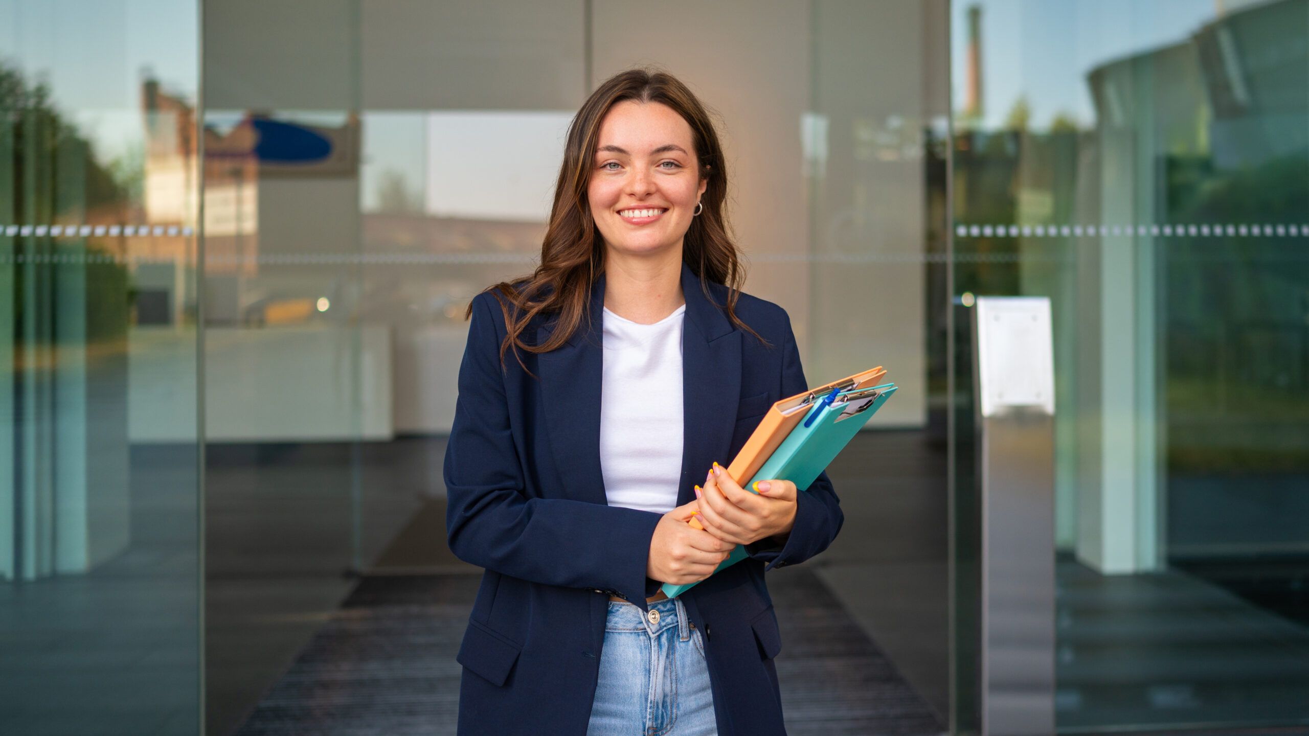 Montaire Student Living Apartments Young woman in a blazer holding folders and smiling, standing in front of glass doors.