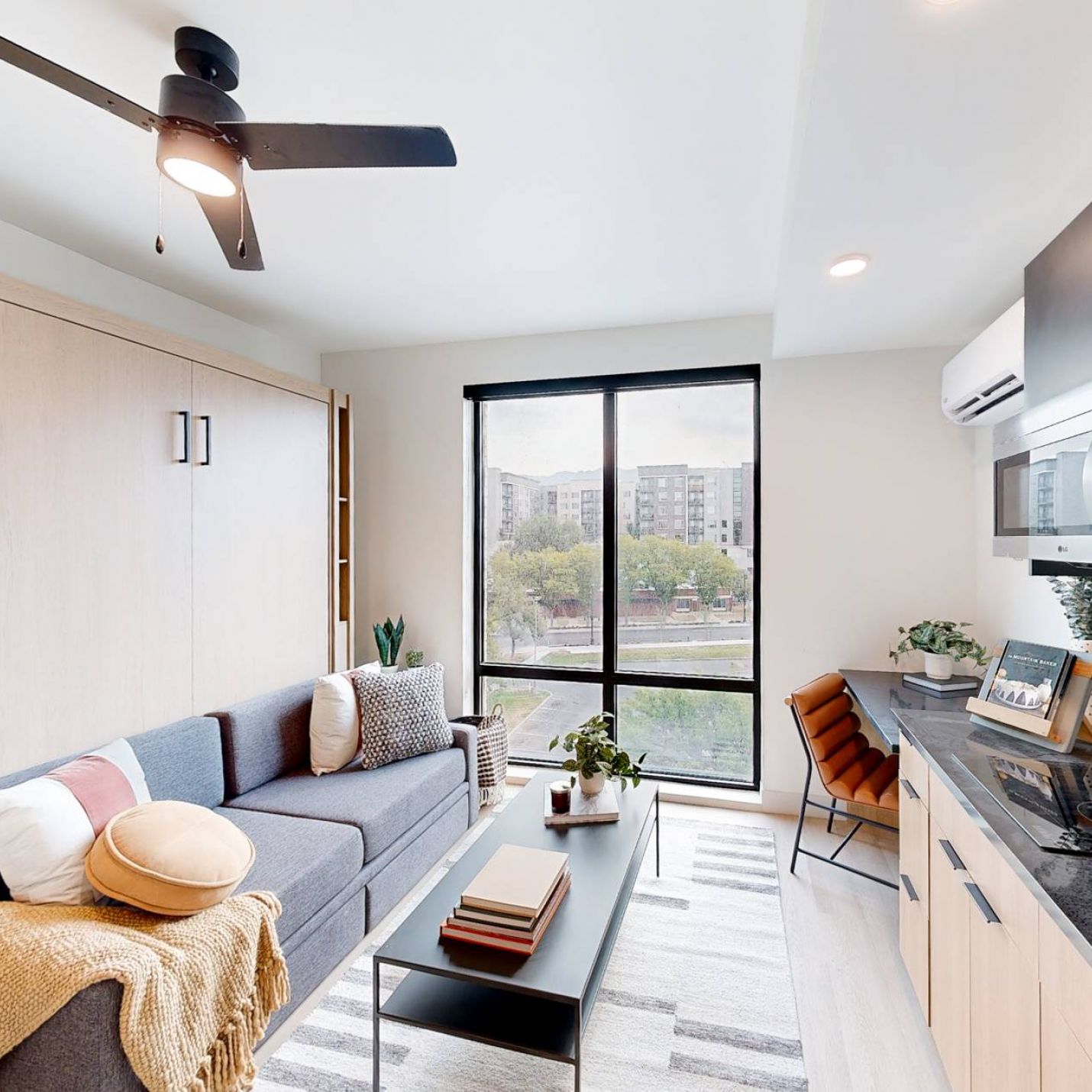 Montaire Student Living Apartments Modern studio apartment with a sofa, kitchen area, and large window overlooking city buildings.