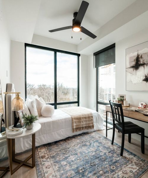 Montaire Student Living Apartments Modern bedroom with large windows, a single bed, a desk, a chair, a mirror, and a ceiling fan.