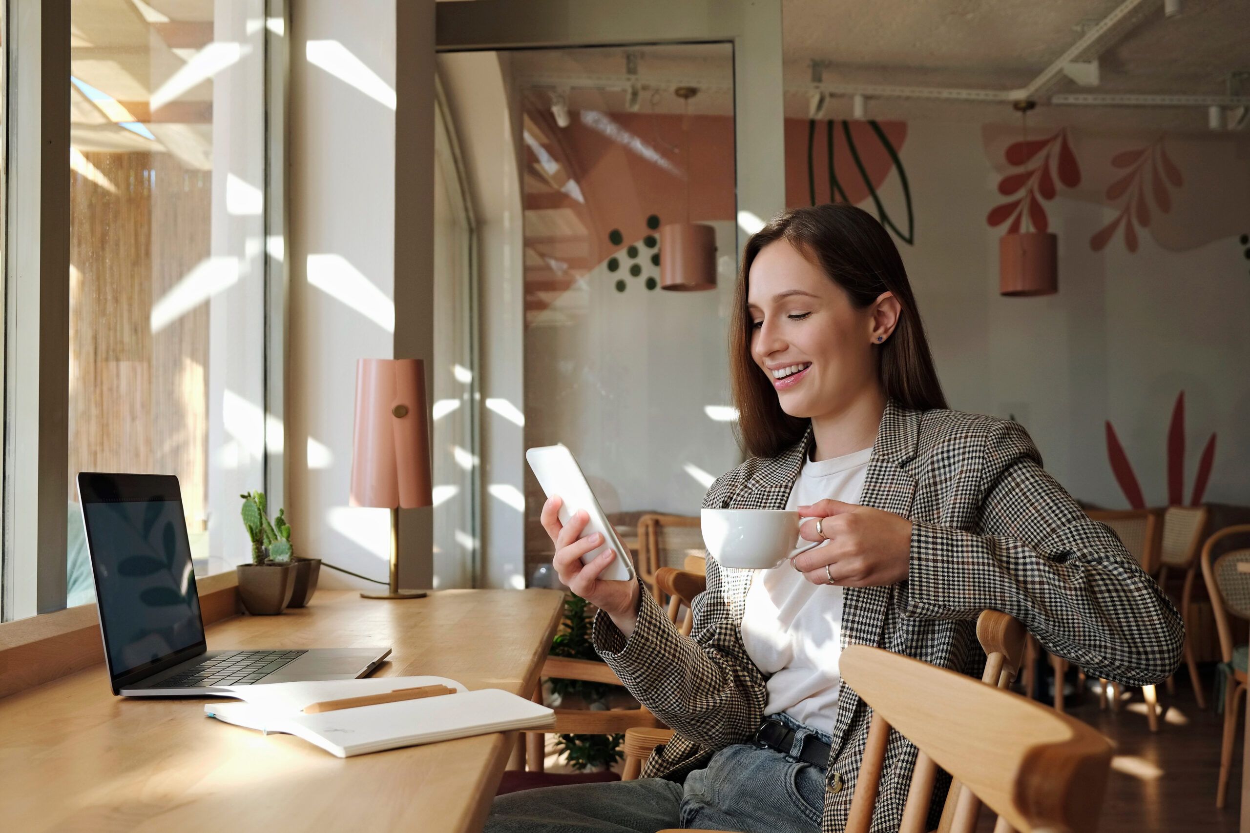 Montaire Student Living Apartments Woman smiling at phone while holding a cup of coffee, sitting at a cafe table with a laptop and notebook.