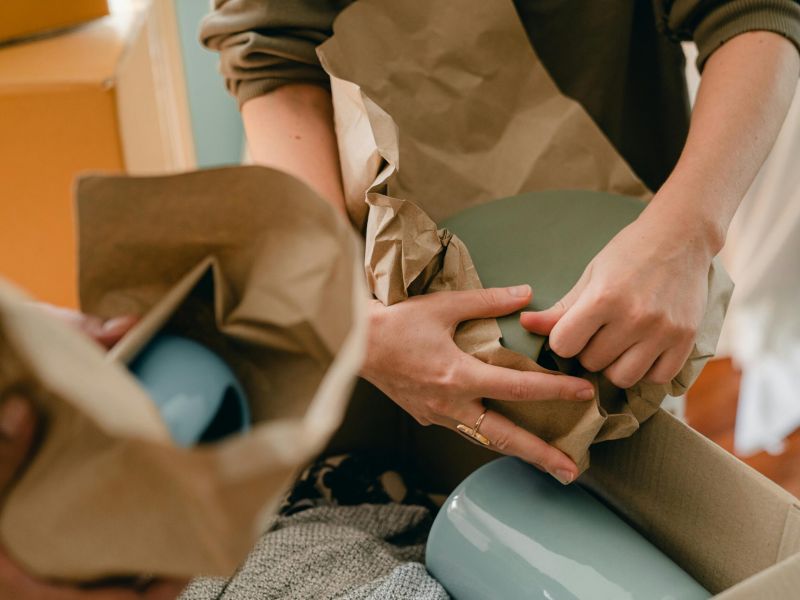 Montaire Student Living Apartments Hands wrapping blue ceramics in brown paper for packing, with boxes and cloth visible in the background.