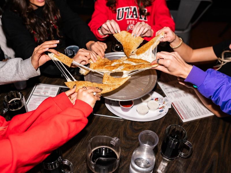 Montaire Student Living Apartments Five people pull cheesy pizza slices from a pizza pan, with drinks and menus on a wooden table.