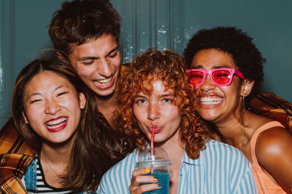 Montaire Student Living Apartments Four friends smiling and laughing together; one drinks a blue beverage with a straw, another wears pink sunglasses.