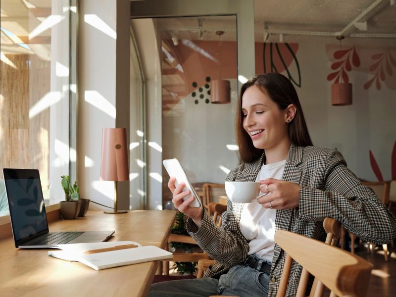 Montaire Student Living Apartments Woman smiling at phone while holding a cup of coffee, sitting at a cafe table with a laptop and notebook.
