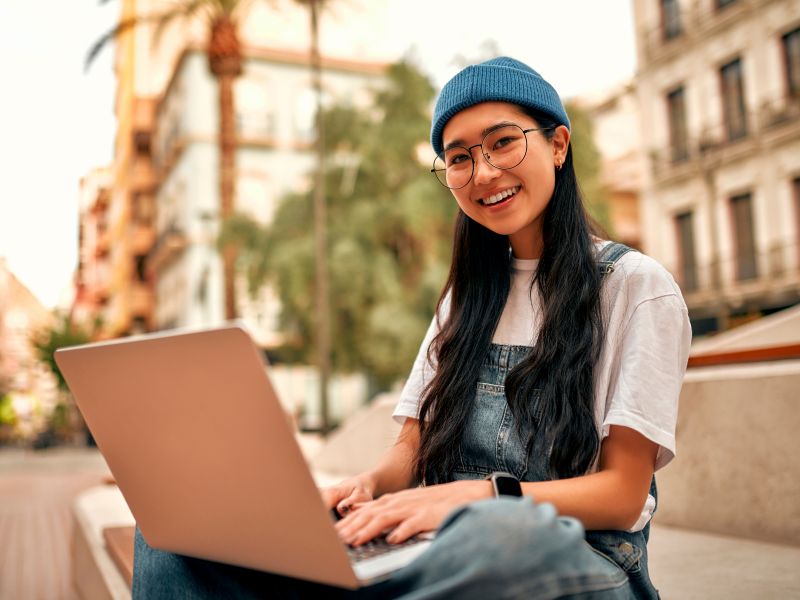 Montaire Student Living Apartments Young woman with long hair and glasses, wearing a beanie, using a laptop outdoors in a city setting, smiling.