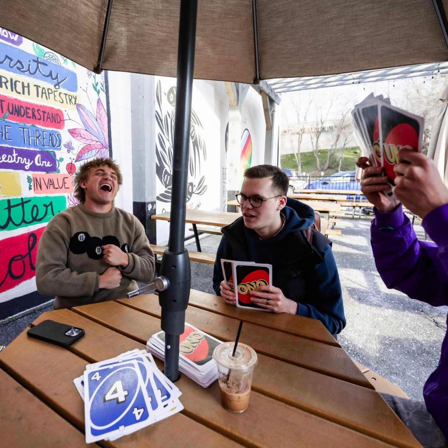 Montaire Student Living Apartments Four friends playing UNO and laughing around a picnic table outdoors, with colorful murals in the background.
