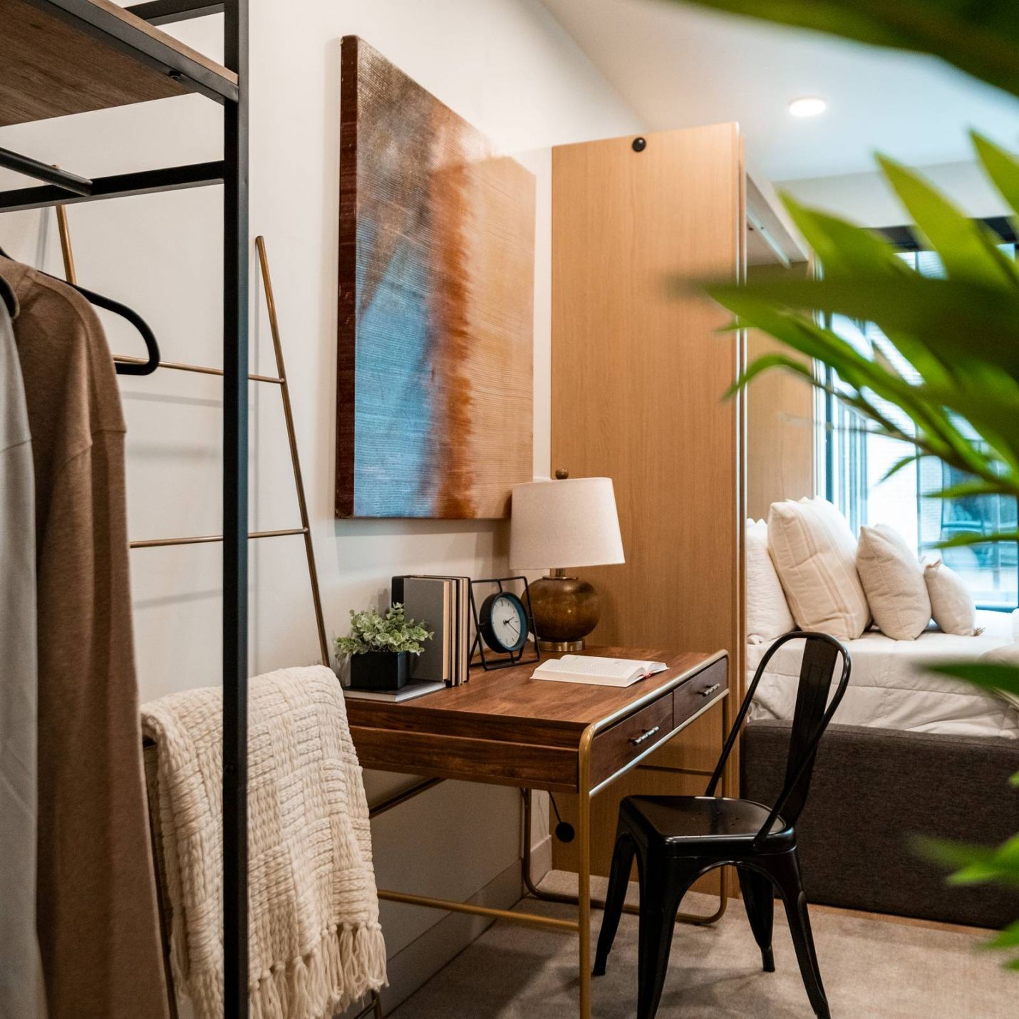 Montaire Student Living Apartments A cozy bedroom with a desk, chair, lamp, clothes rack, and bed, with greenery in the foreground.