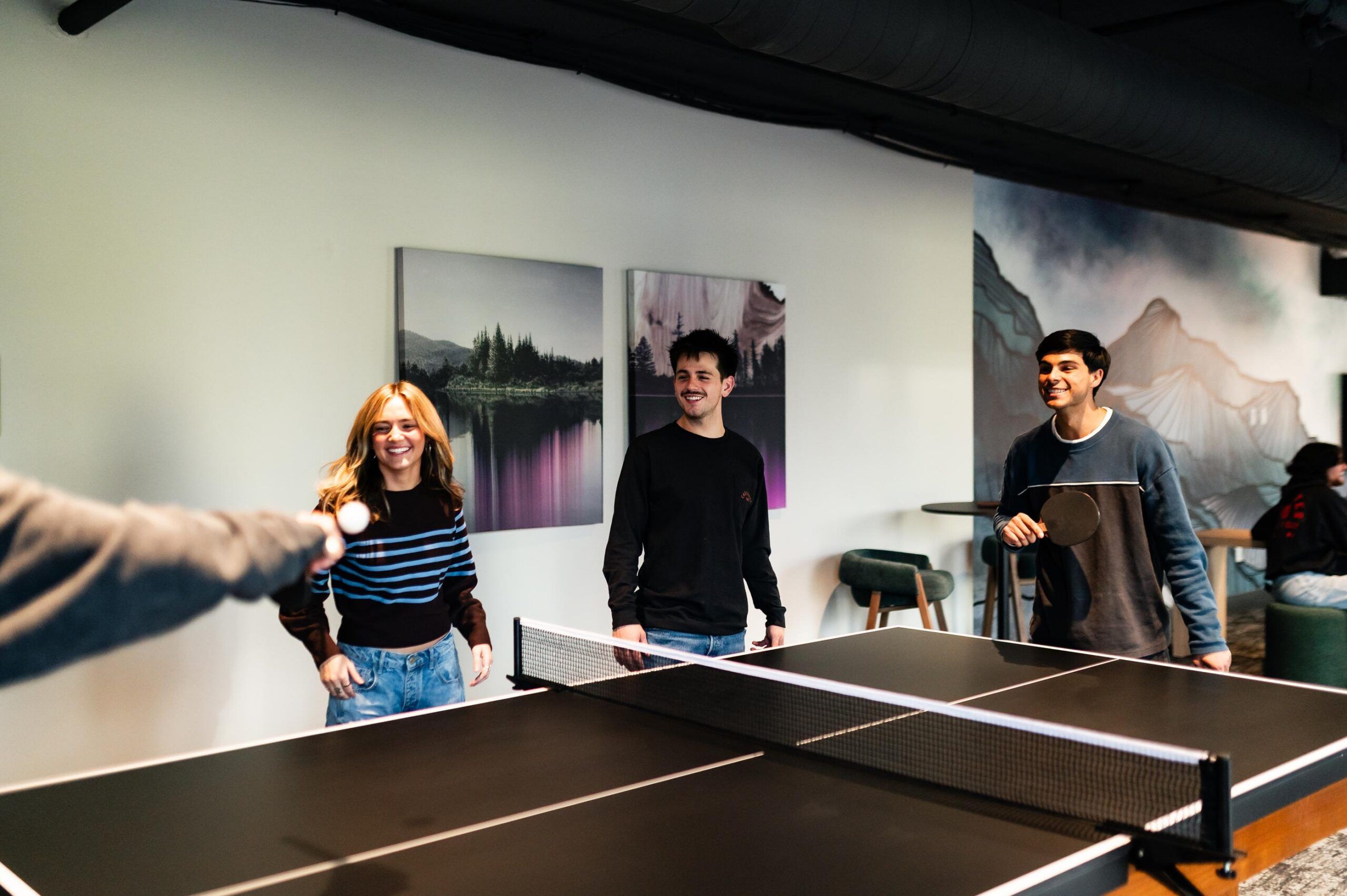 Montaire Student Living Apartments Three young adults smile and play table tennis indoors, two holding paddles, artwork visible on the wall behind them.