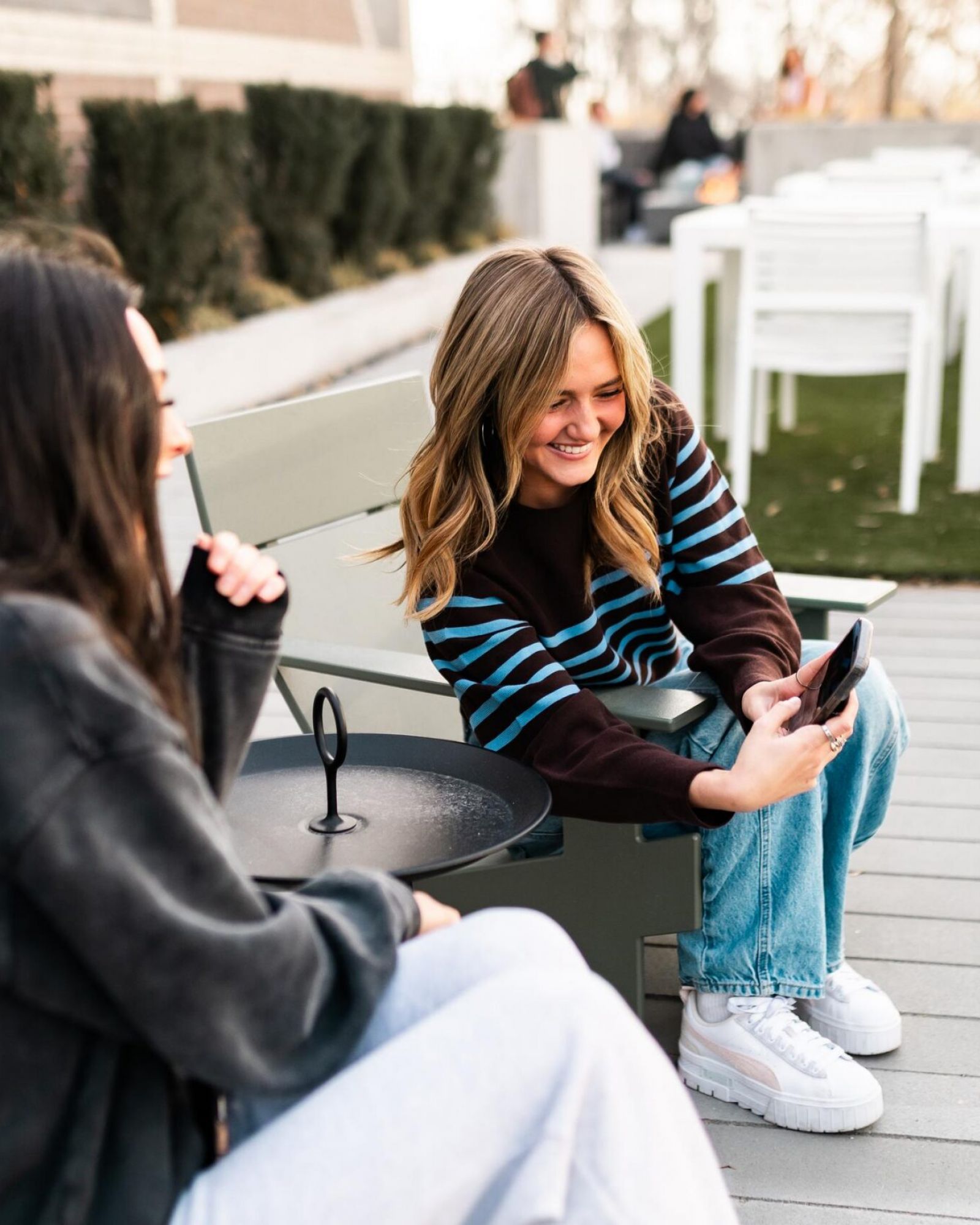 Montaire Student Living Apartments Two women sit outside on a patio bench, smiling and looking at a phone together on a sunny day.