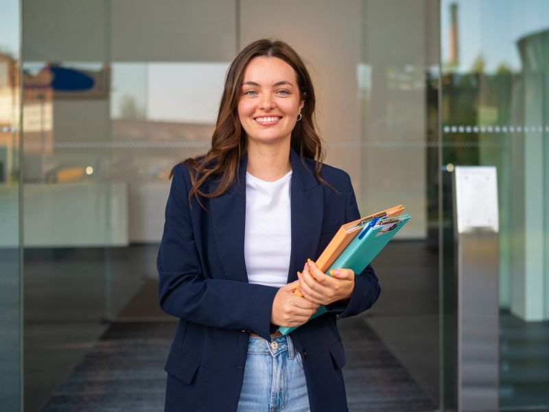 Montaire Student Living Apartments Young woman in a blazer holding folders and smiling, standing in front of glass doors.