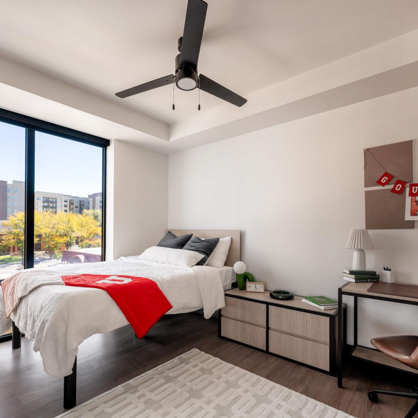 Montaire Student Living Apartments A modern bedroom featuring a bed with a red throw blanket, a desk with a brown chair, and a view of an urban landscape through a large window.