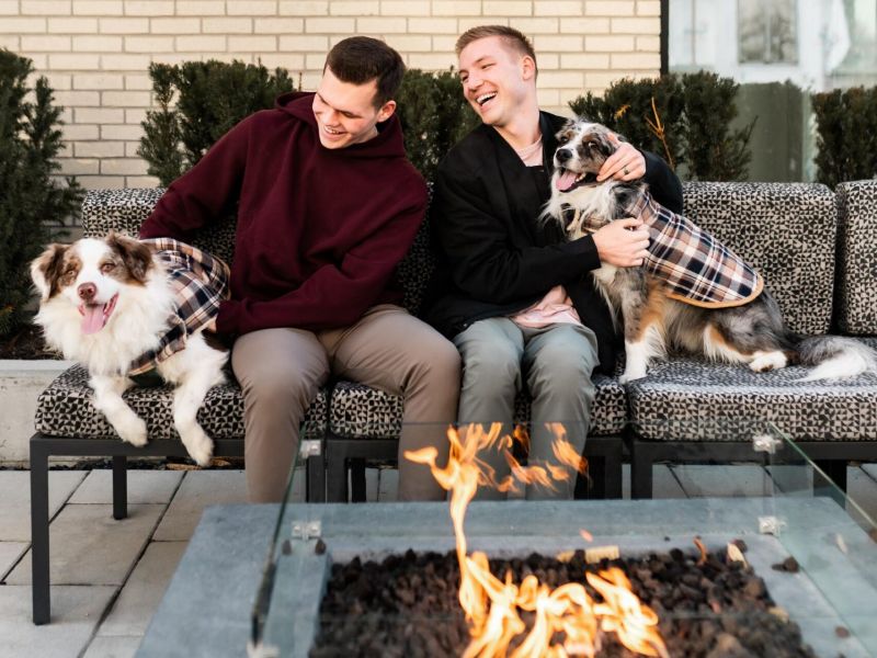 Montaire Student Living Apartments Two men on an outdoor bench laugh with their two dogs in plaid coats beside a modern fire pit.