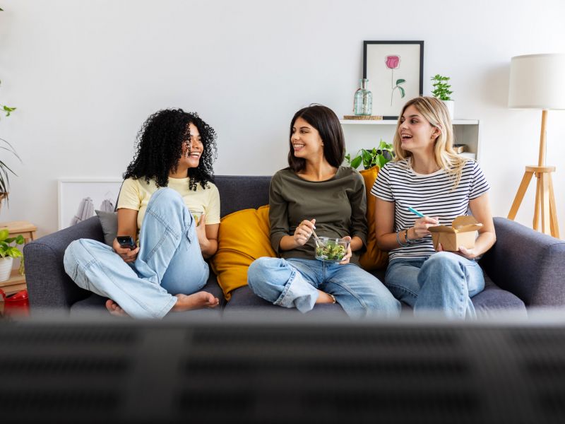 Montaire Student Living Apartments Three women sit on a couch, eating and smiling, watching TV in a cozy, plant-filled living room.