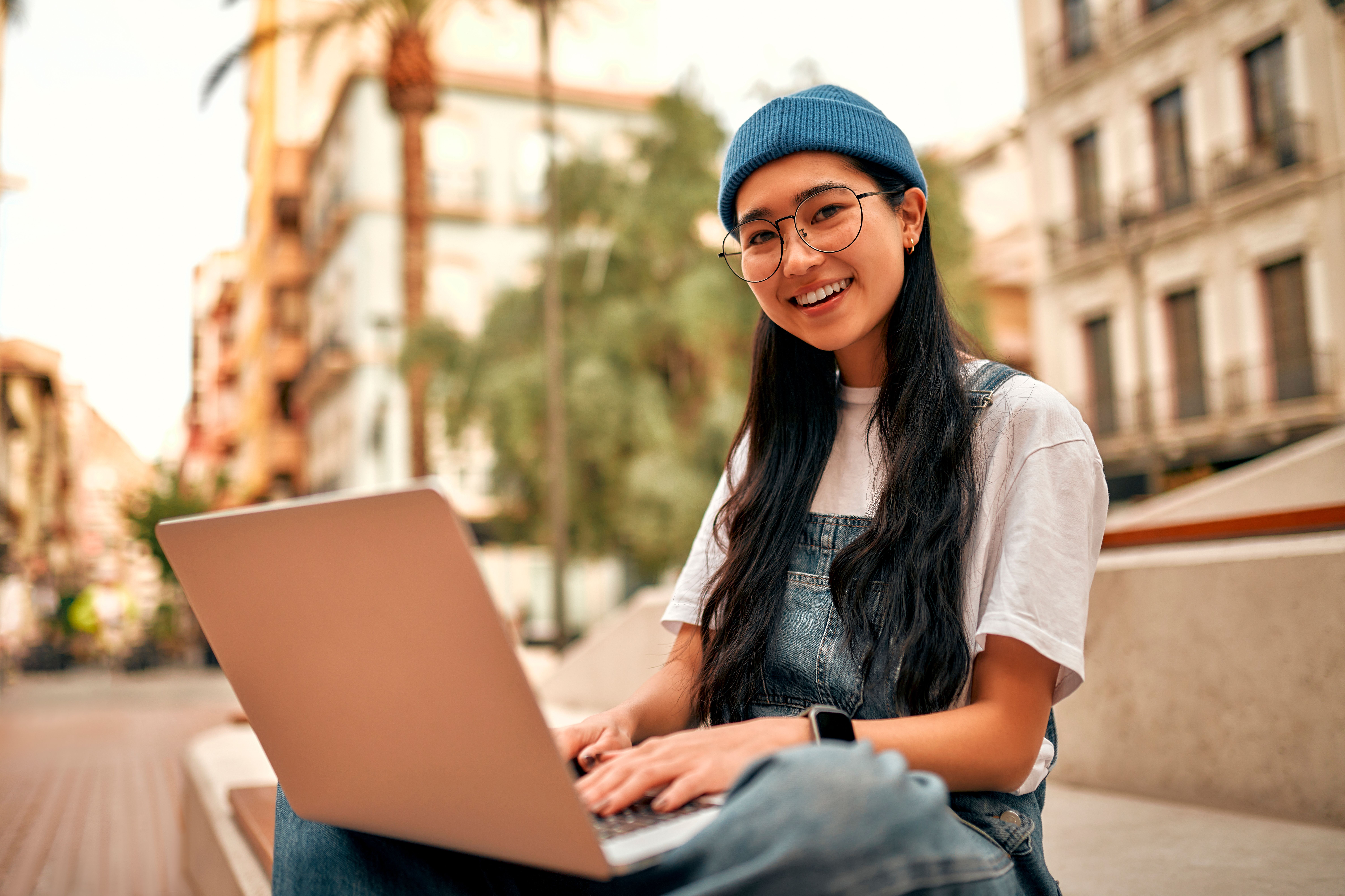 Montaire Student Living Apartments Young woman with long hair and glasses, wearing a beanie, using a laptop outdoors in a city setting, smiling.