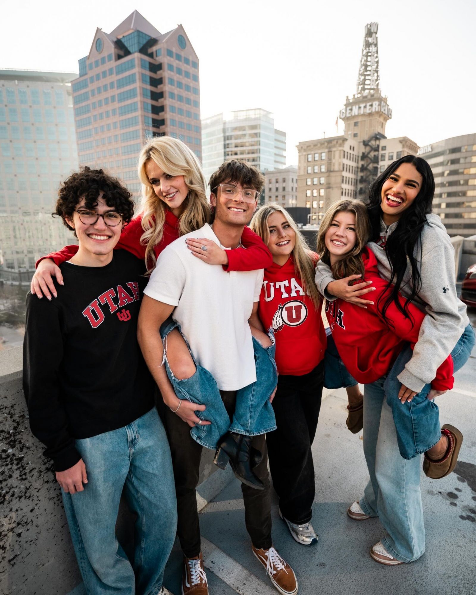 Montaire Student Living Apartments Six smiling young adults in Utah apparel pose together on a rooftop with city buildings in the background.