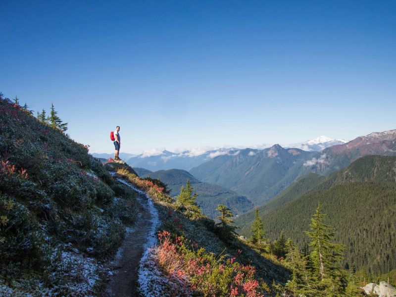 Montaire Student Living Apartments A hiker stands on a mountain trail, overlooking forested hills and distant snowy peaks under a clear blue sky.