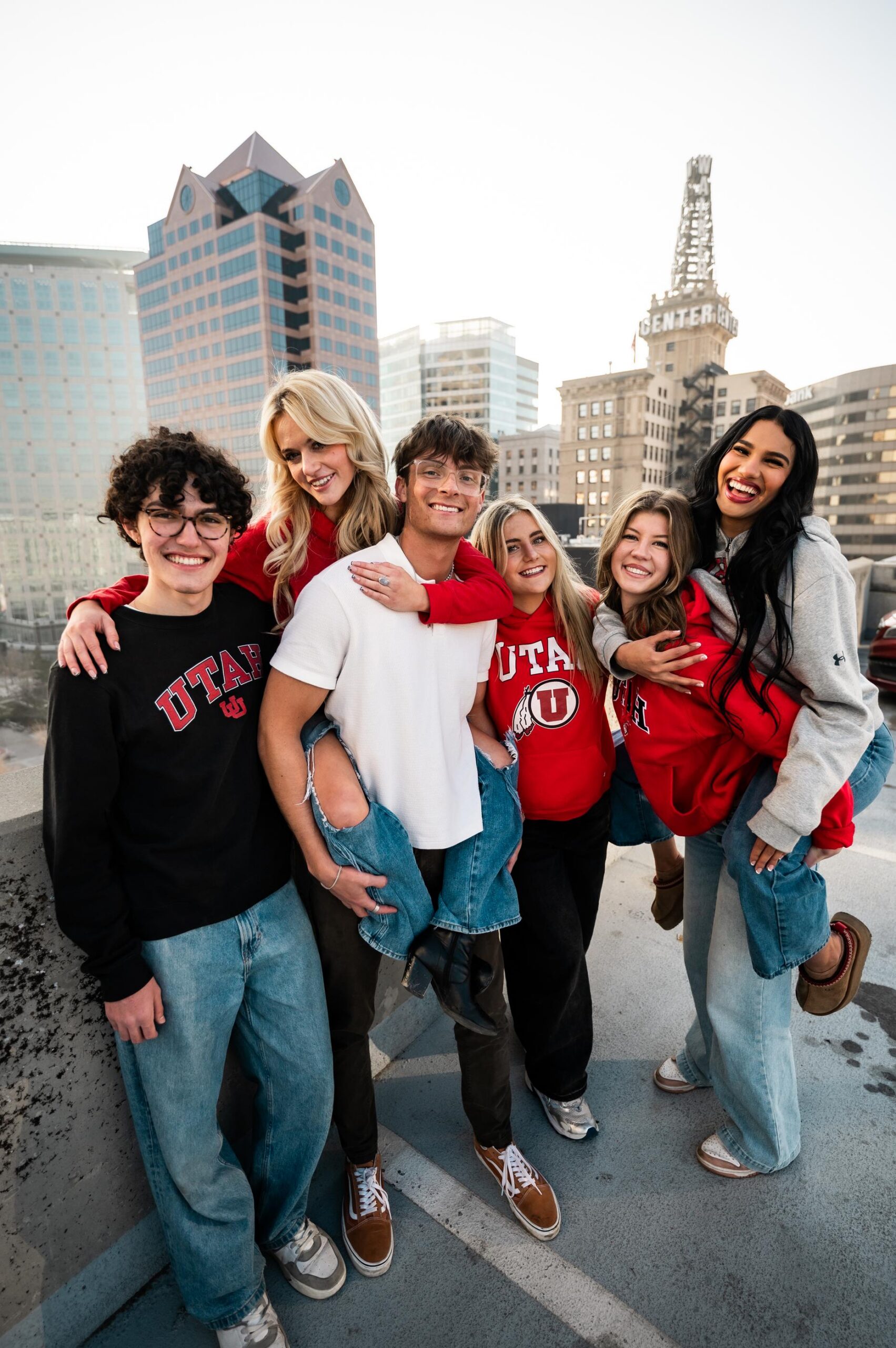 Montaire Student Living Apartments Six smiling young adults in Utah apparel pose together on a rooftop with city buildings in the background.