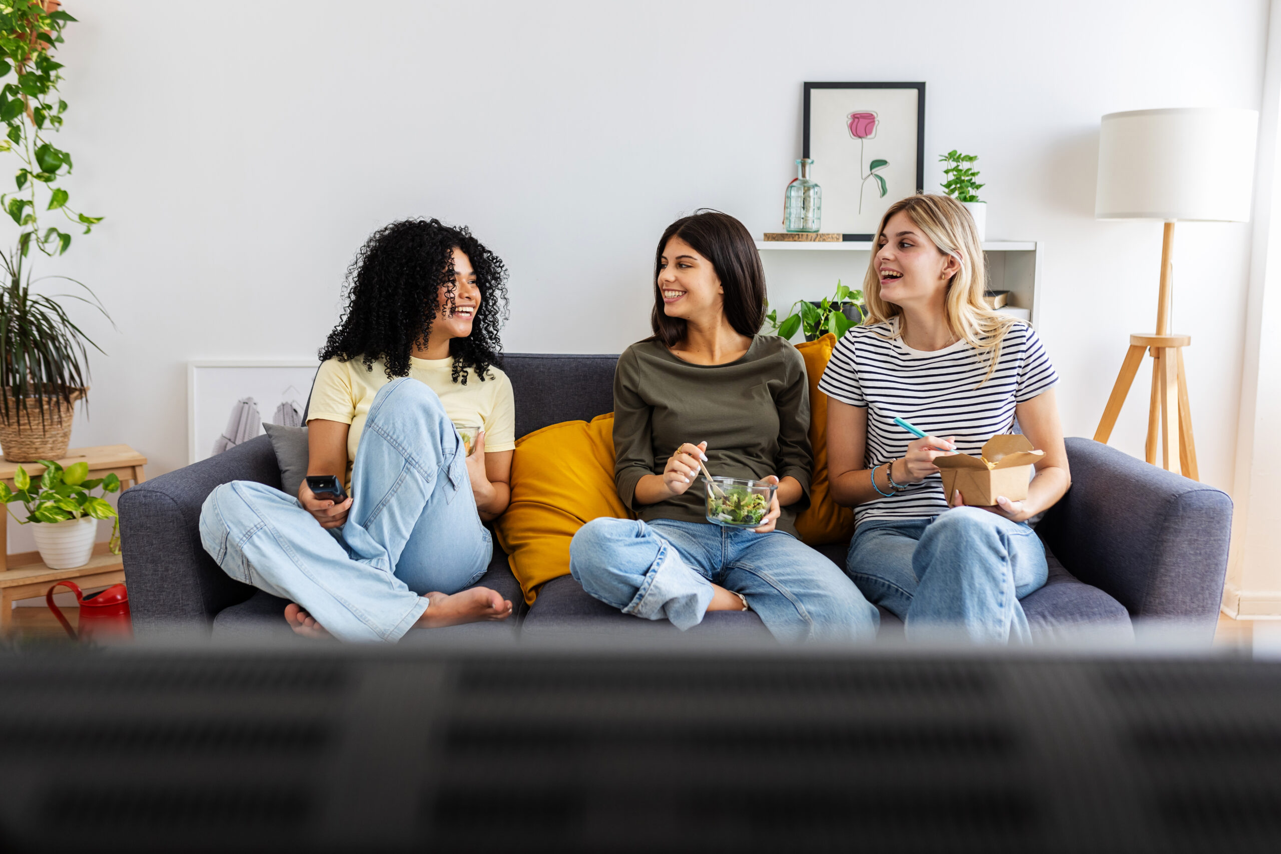 Montaire Student Living Apartments Three women sit on a couch, eating and smiling, watching TV in a cozy, plant-filled living room.