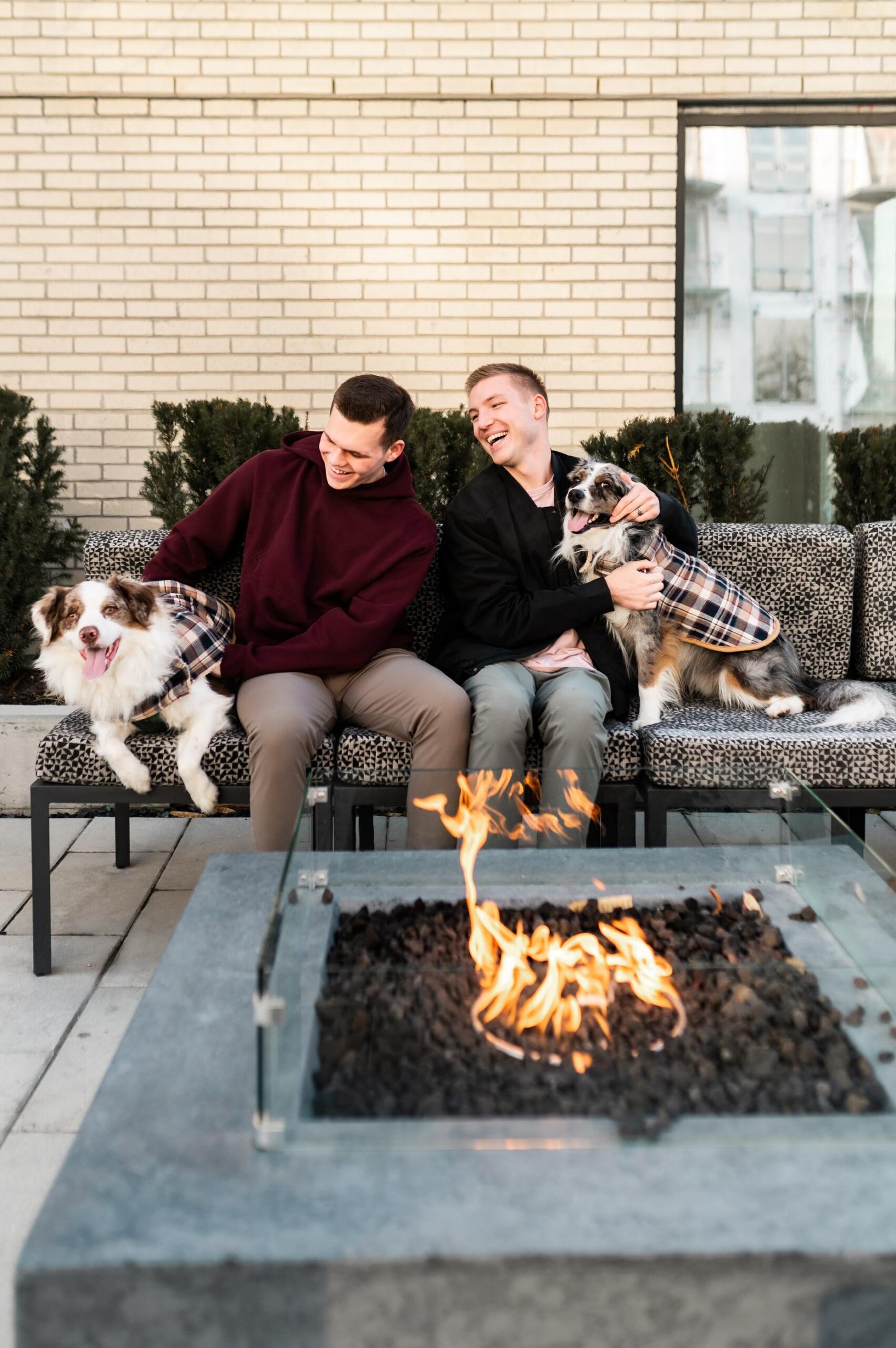 Montaire Student Living Apartments Two men on an outdoor bench laugh with their two dogs in plaid coats beside a modern fire pit.