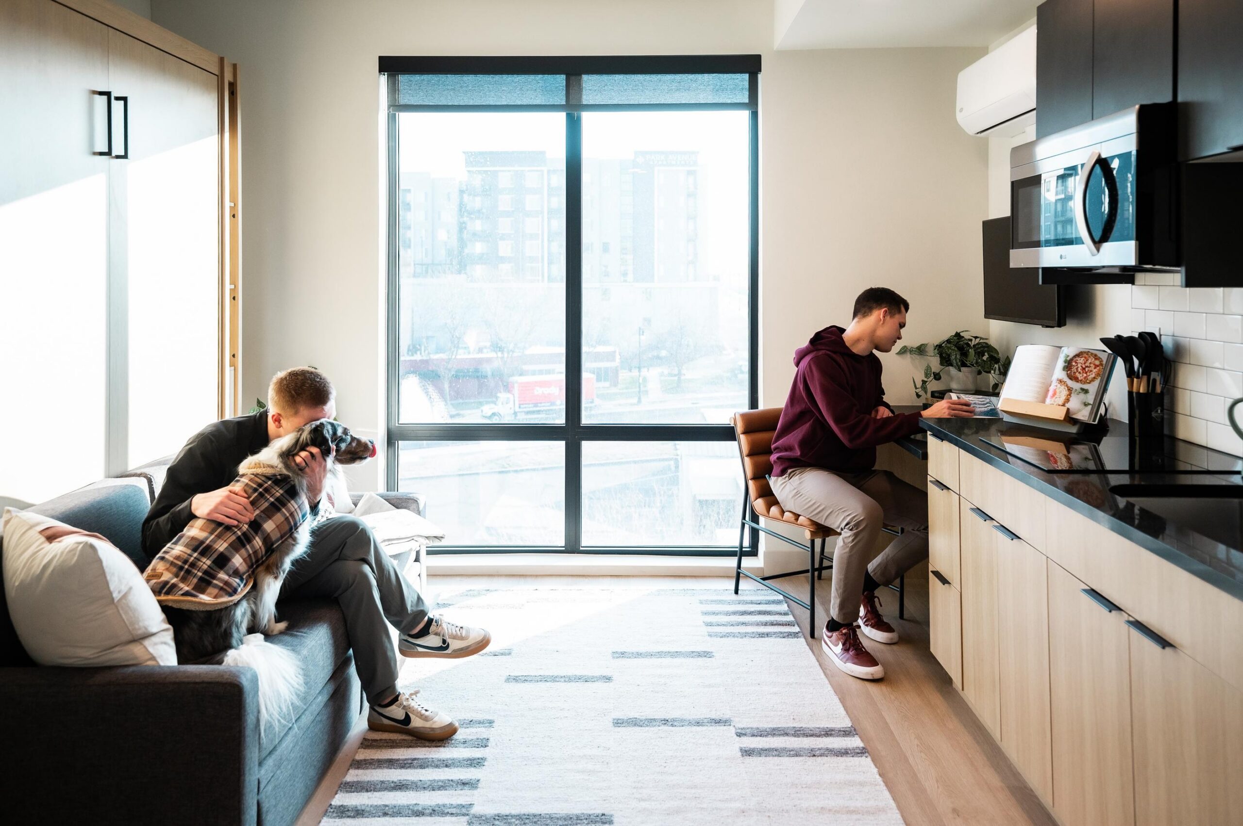 Montaire Student Living Apartments Two people in a modern apartment, one working at a kitchen counter, the other cuddling a dog on the couch.